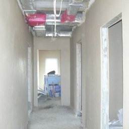 PROBUD ST SPÓŁKA Z OGRANICZONĄ ODPOWIEDZIALNOŚCIĄ - Korytarz w trakcie prac wykończeniowych: świeżo położone tynki na ścianach, widoczne instalacje pod sufitem, przygotowane ościeżnice. W głębi pomieszczenia materiały budowlane.