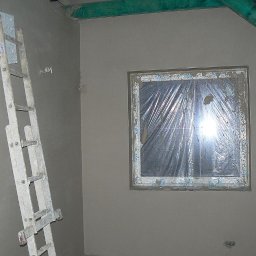 PROBUD ST SPÓŁKA Z OGRANICZONĄ ODPOWIEDZIALNOŚCIĄ - Ściana po nałożeniu tynku, z oknem zabezpieczonym folią i drabiną opartą o ścianę. Widoczne zielone elementy konstrukcji dachu.