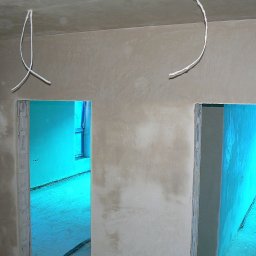 PROBUD ST SPÓŁKA Z OGRANICZONĄ ODPOWIEDZIALNOŚCIĄ - Wnętrze w trakcie prac wykończeniowych: ściany po szpachlowaniu, widoczne kable zwisające z sufitu, przygotowane otwory drzwiowe. W tle okno z niebieską folią ochronną.