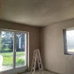 PROBUD ST SPÓŁKA Z OGRANICZONĄ ODPOWIEDZIALNOŚCIĄ - Pokój po tynkowaniu maszynowym: gładkie, jasne ściany, okno z widokiem na zieleń, drabina malarska w rogu. Świeżo nałożony tynk, gotowy do dalszych prac wykończeniowych.