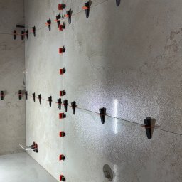 PROBUD ST SPÓŁKA Z OGRANICZONĄ ODPOWIEDZIALNOŚCIĄ - Ściana i podłoga w trakcie układania płytek, z użyciem systemu poziomowania płytek (klipsy i kliny) oraz sznurka wyznaczającego linię. Widoczne detale faktury płytek.