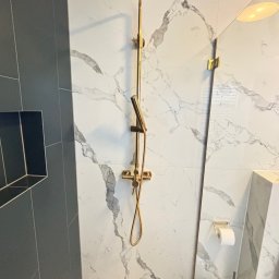 PROBUD ST SPÓŁKA Z OGRANICZONĄ ODPOWIEDZIALNOŚCIĄ - Nowoczesna łazienka z kabiną typu walk-in, wykończona płytkami imitującymi marmur i złotą armaturą, w tym deszczownicą i słuchawką prysznicową.