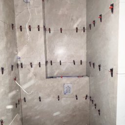 PROBUD ST SPÓŁKA Z OGRANICZONĄ ODPOWIEDZIALNOŚCIĄ - Ściany łazienki wyłożone płytkami w trakcie montażu, z użyciem systemu poziomowania płytek. Widoczne wiaderka i fragment płytki na podłodze.