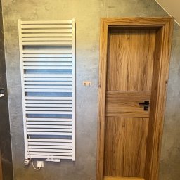 PROBUD ST SPÓŁKA Z OGRANICZONĄ ODPOWIEDZIALNOŚCIĄ - Wykończona łazienka z grzejnikiem drabinkowym, drewnianą podłogą i drzwiami z drewnianą ościeżnicą. Szare płytki na ścianach. Ukośny sufit.