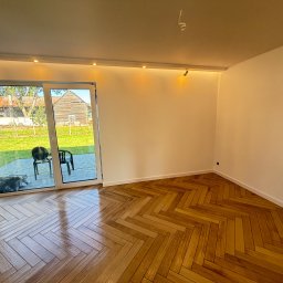 PROBUD ST SPÓŁKA Z OGRANICZONĄ ODPOWIEDZIALNOŚCIĄ - Jasne, odnowione wnętrze z parkietem w jodełkę, białymi ścianami i dużym oknem tarasowym z widokiem na zieleń. Nowoczesne oświetlenie sufitowe.