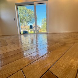 PROBUD ST SPÓŁKA Z OGRANICZONĄ ODPOWIEDZIALNOŚCIĄ - Połyskująca, drewnopodobna podłoga ułożona w jodełkę, z widokiem na zieleń przez przesuwne drzwi balkonowe. Minimalistyczne wnętrze z jasnymi ścianami.