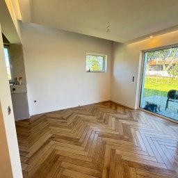 PROBUD ST SPÓŁKA Z OGRANICZONĄ ODPOWIEDZIALNOŚCIĄ - Jasne, parkietowe panele ułożone w jodełkę w przestronnym, odnowionym pokoju z dużym oknem tarasowym i świeżo pomalowanymi ścianami. Widok na zieleń za oknem.