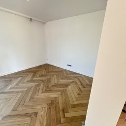 PROBUD ST SPÓŁKA Z OGRANICZONĄ ODPOWIEDZIALNOŚCIĄ - Jasne, puste wnętrze z parkietem ułożonym w jodełkę. Świeżo pomalowane ściany i listwy przypodłogowe. Widoczny fragment kolumny po prawej stronie kadru.