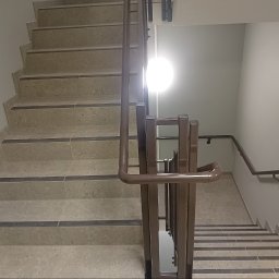PROBUD ST SPÓŁKA Z OGRANICZONĄ ODPOWIEDZIALNOŚCIĄ - Schody z jasnych płytek kamiennych z ciemnymi paskami antypoślizgowymi i brązową metalową balustradą, oświetlone punktowym światłem.