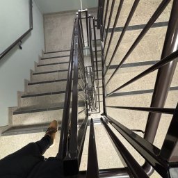 PROBUD ST SPÓŁKA Z OGRANICZONĄ ODPOWIEDZIALNOŚCIĄ - Widok z góry na klatkę schodową z beżowymi płytkami, czarnymi pasami antypoślizgowymi i ciemną, metalową balustradą. W lewym dolnym rogu widoczna stopa w brązowym bucie.