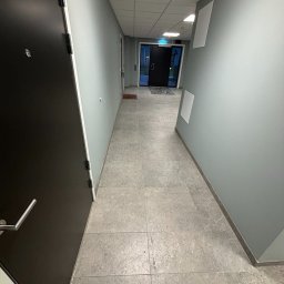PROBUD ST SPÓŁKA Z OGRANICZONĄ ODPOWIEDZIALNOŚCIĄ - Nowoczesny, długi korytarz z jasnoszarymi, gładkimi ścianami i szarą podłogą z dużych płytek. Po lewej ciemne drzwi z metalową klamką, w tle przeszklone wyjście. Sufit podwieszany z oświetleniem...