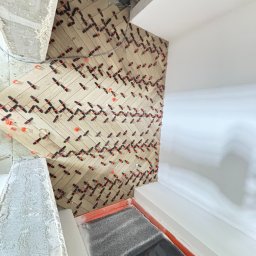 PROBUD ST SPÓŁKA Z OGRANICZONĄ ODPOWIEDZIALNOŚCIĄ - Układanie płytek imitujących drewno w jodełkę na ścianie, z licznymi czarno-pomarańczowymi krzyżykami dystansowymi. Widoczny surowy betonowy element i gładka, biała ściana.