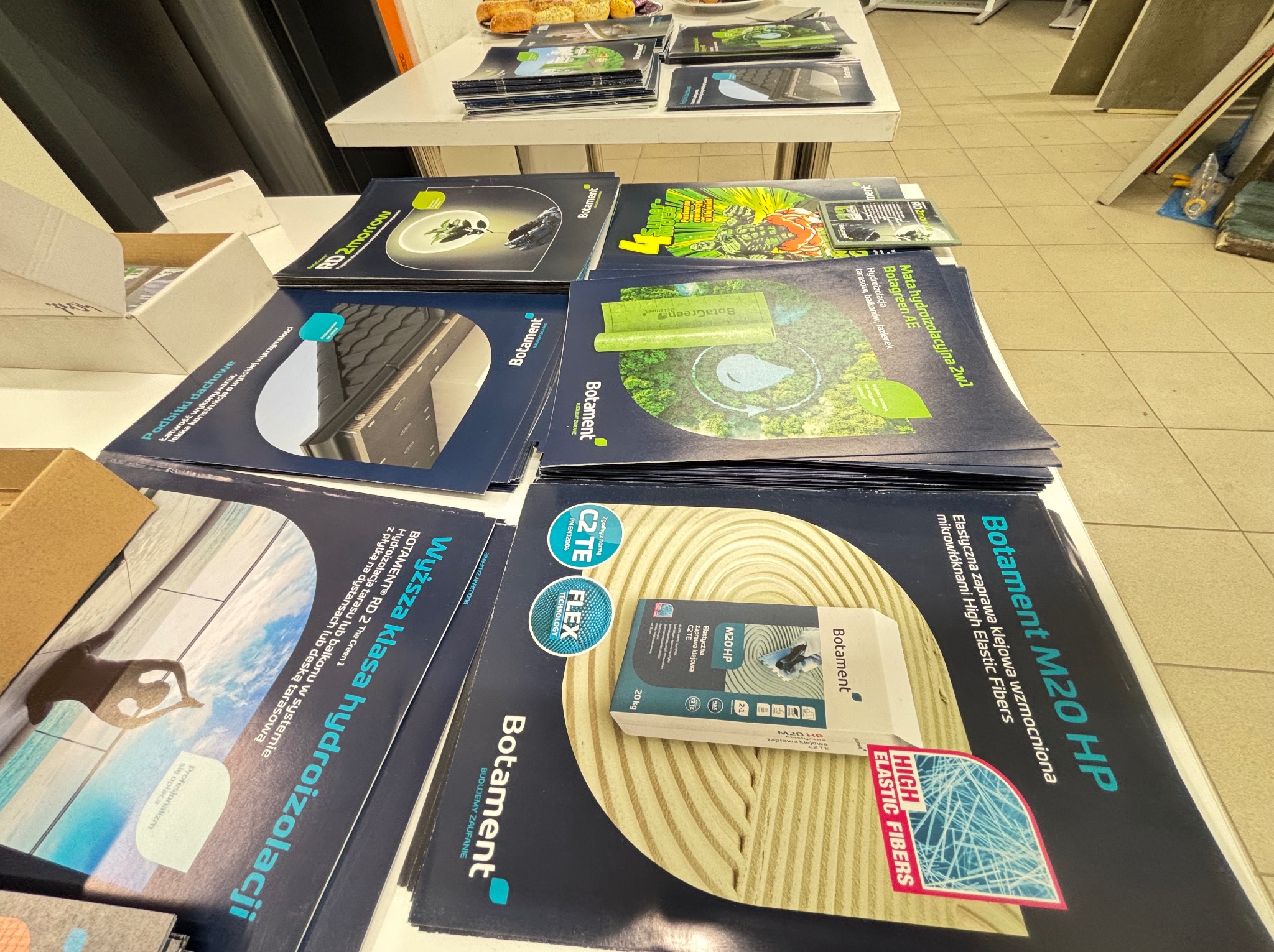 Stos materiałów reklamowych Botament z zakresu hydroizolacji i zapraw klejowych na stole. Broszury, ulotki i próbki produktów. Widoczne hasła: hydroizolacja, elastyczna zaprawa klejowa.