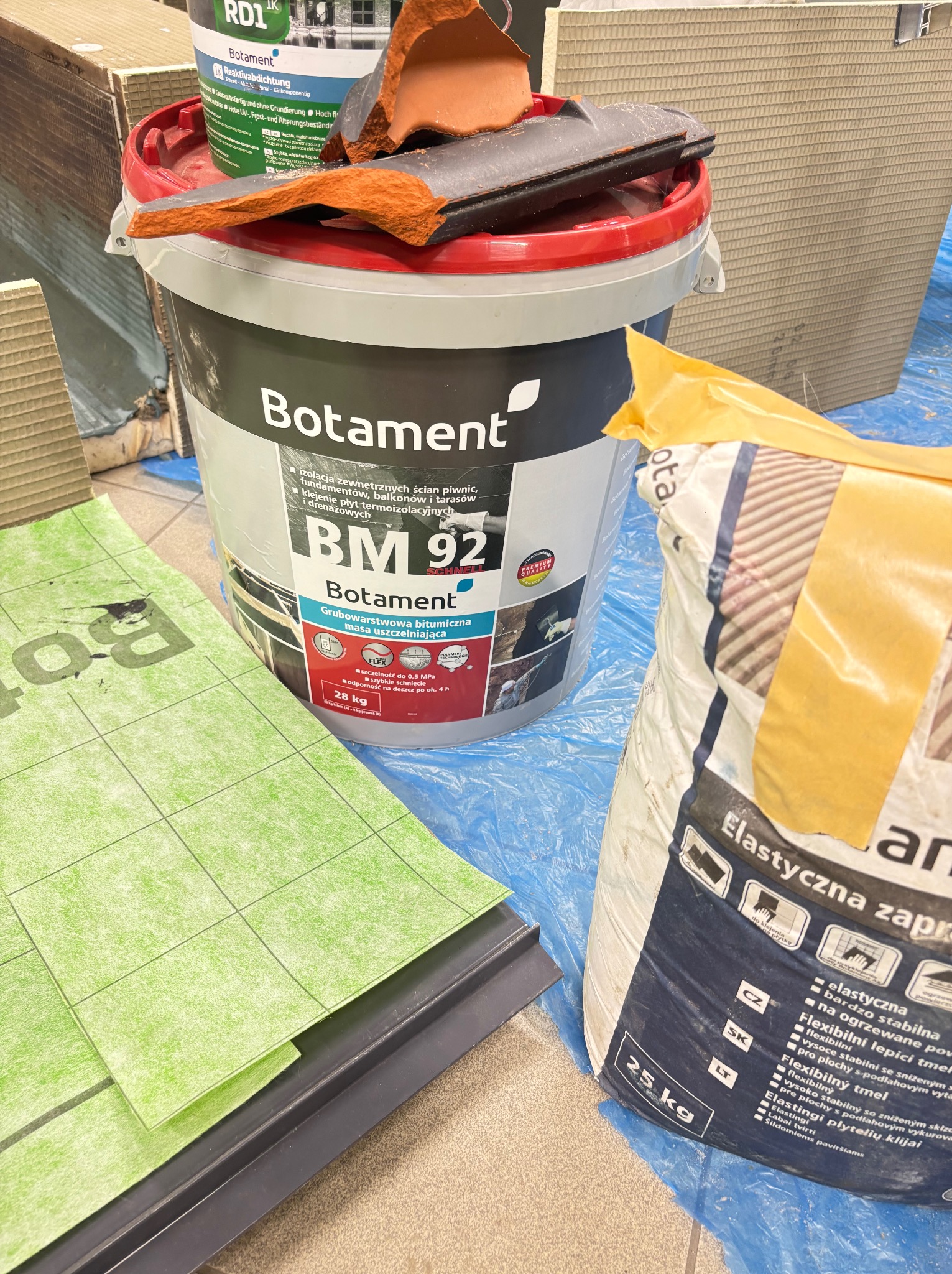 Wiadro z masą bitumiczną Botament BM 92, obok zielona mata uszczelniająca i worek elastycznej zaprawy klejowej. Fragmenty dachówki na wiadrze. Niebieska folia ochronna.