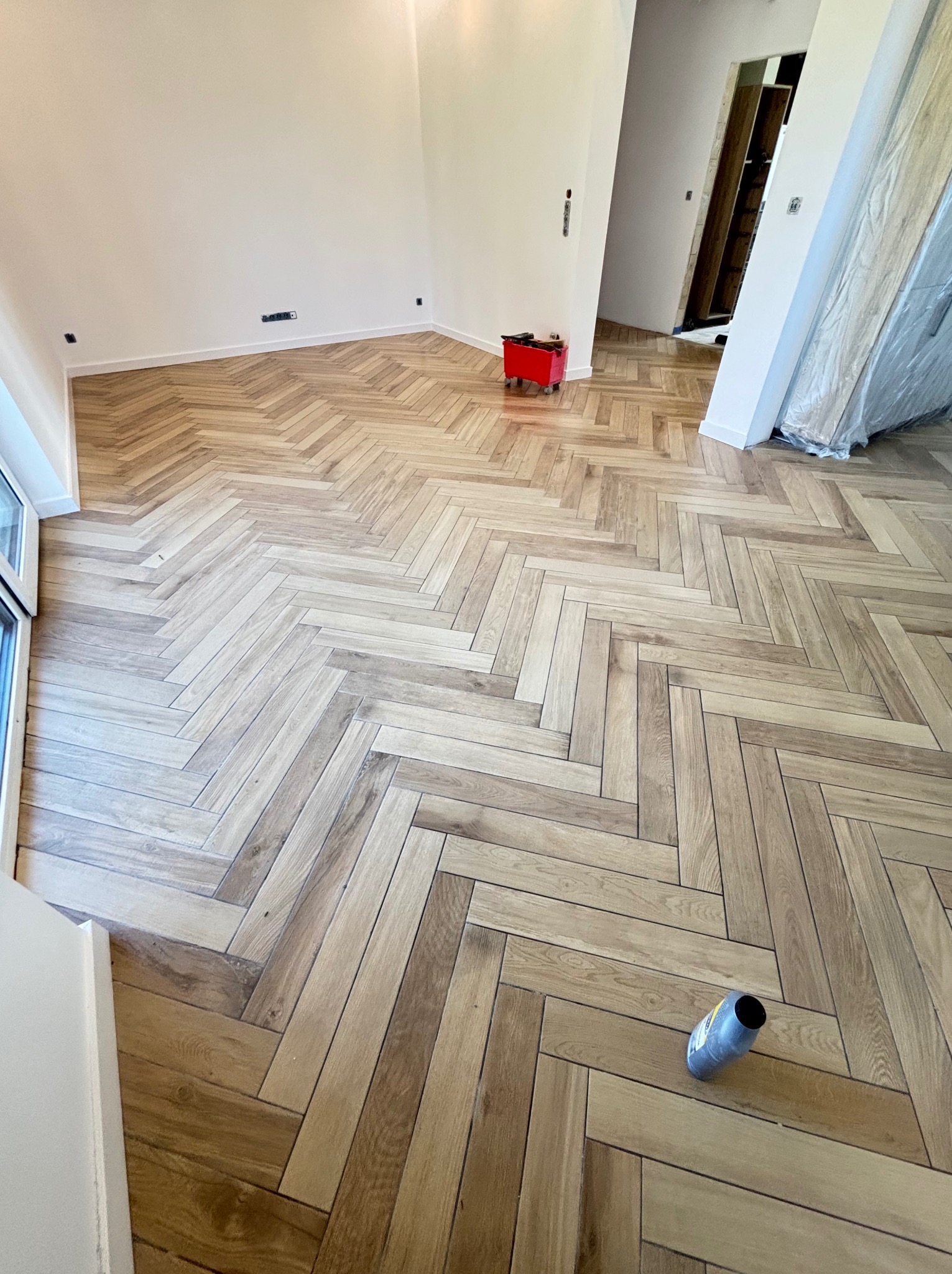 Parkiet ułożony w jodełkę z płytek imitujących drewno w jasnym wnętrzu. Widoczny czerwony wózek narzędziowy i butelka po kleju. Nowoczesne wykończenie podłogi.