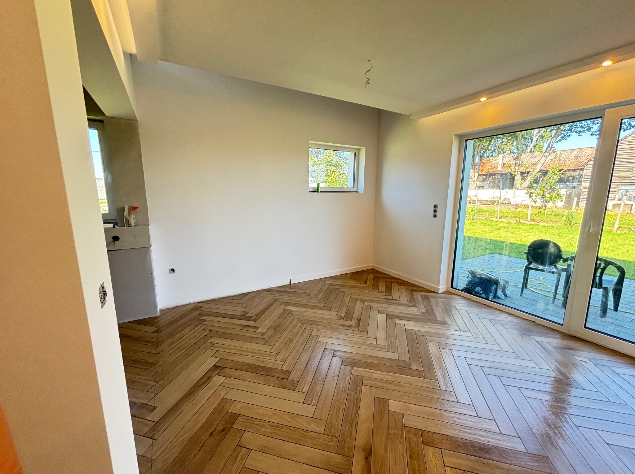 Wykończone wnętrze z parkietem ułożonym w jodełkę, świeżo pomalowane ściany na biało, widok na ogród przez duże okno przesuwne. Nowoczesne wykończenie.