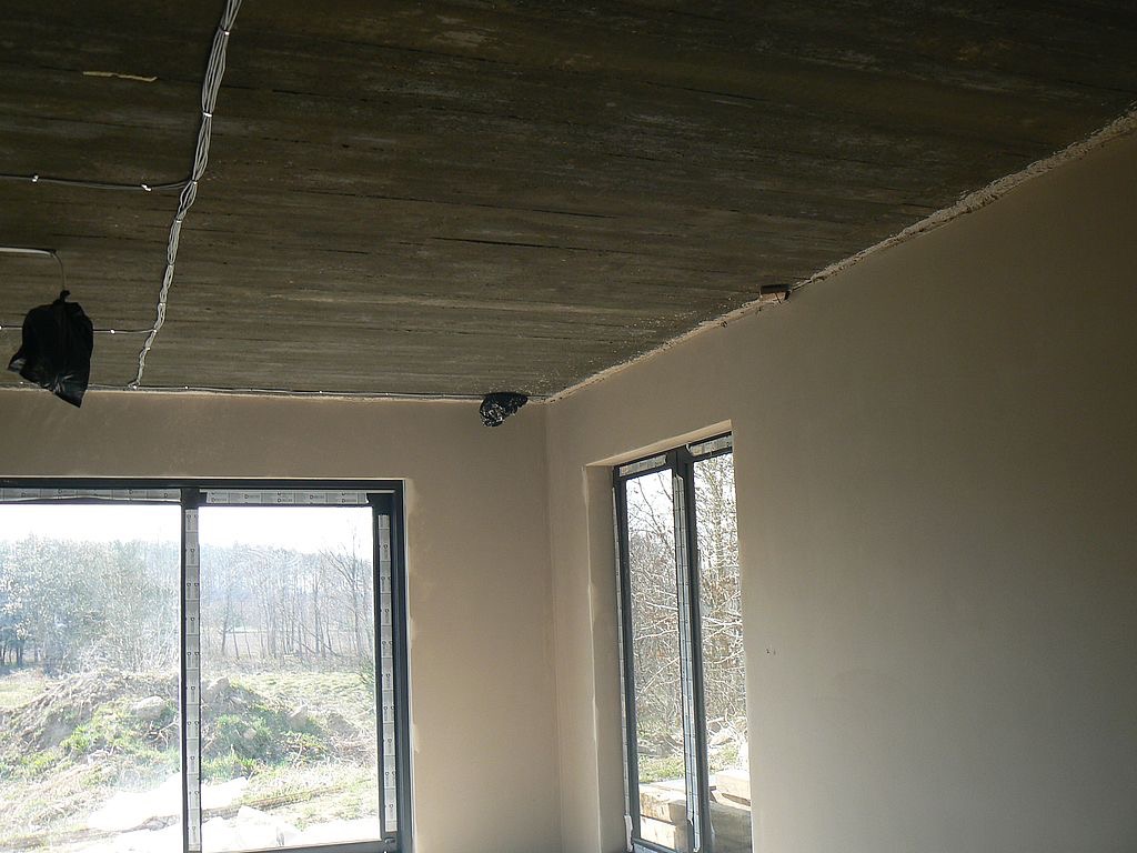 Wnętrze po szpachlowaniu w Sierakowicach. Widoczne gładkie ściany, surowy betonowy sufit z instalacją elektryczną i dwa okna z widokiem na zewnątrz. Jasne, naturalne światło.