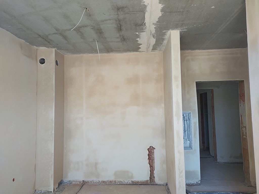 Surowe wnętrze w trakcie prac wykończeniowych: ściany po szpachlowaniu, widoczne przewody elektryczne w suficie, przygotowane do dalszych prac.