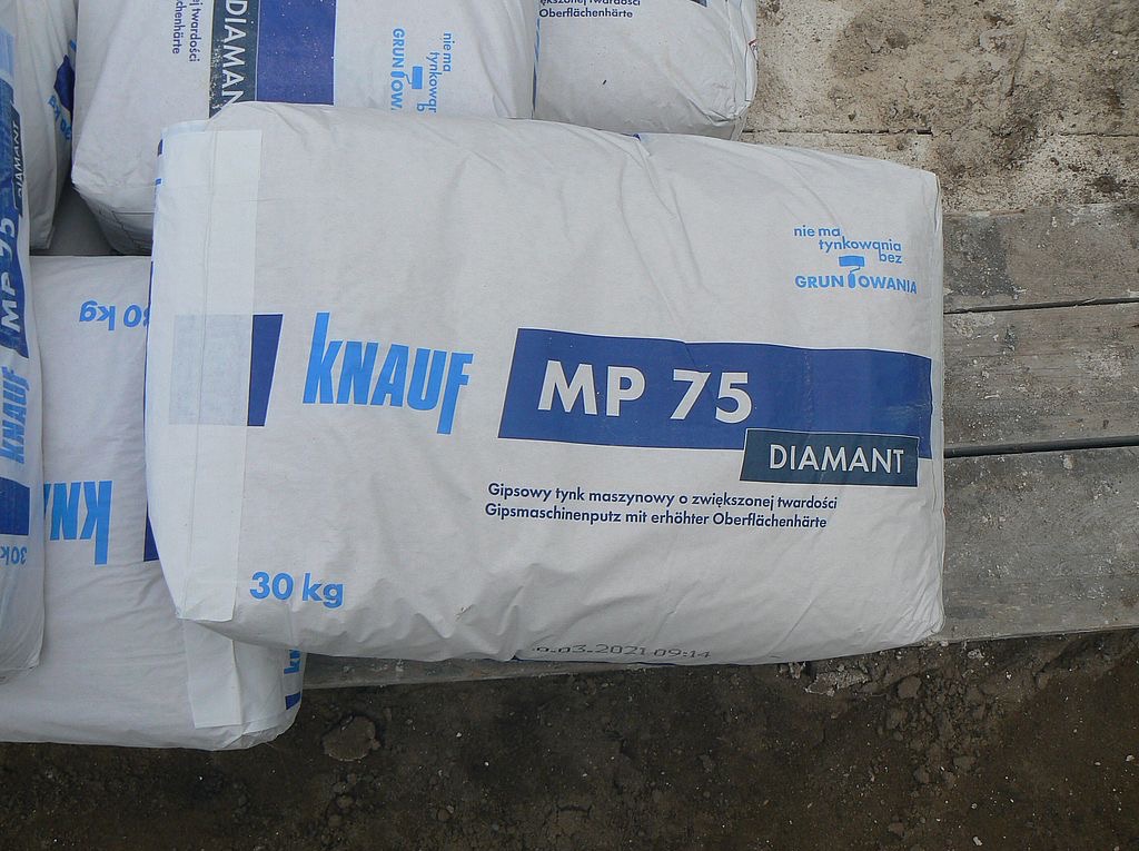 Worek 30 kg gipsowego tynku maszynowego Knauf MP 75 Diamant o zwiększonej twardości, leżący na drewnianej powierzchni. Widoczne inne worki w tle.