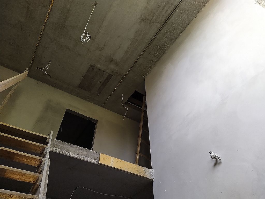 Surowe wnętrze w trakcie prac wykończeniowych: gładka, jasna ściana kontrastuje z surowym betonowym sufitem i drewnianą konstrukcją schodów. Widoczne przewody elektryczne.