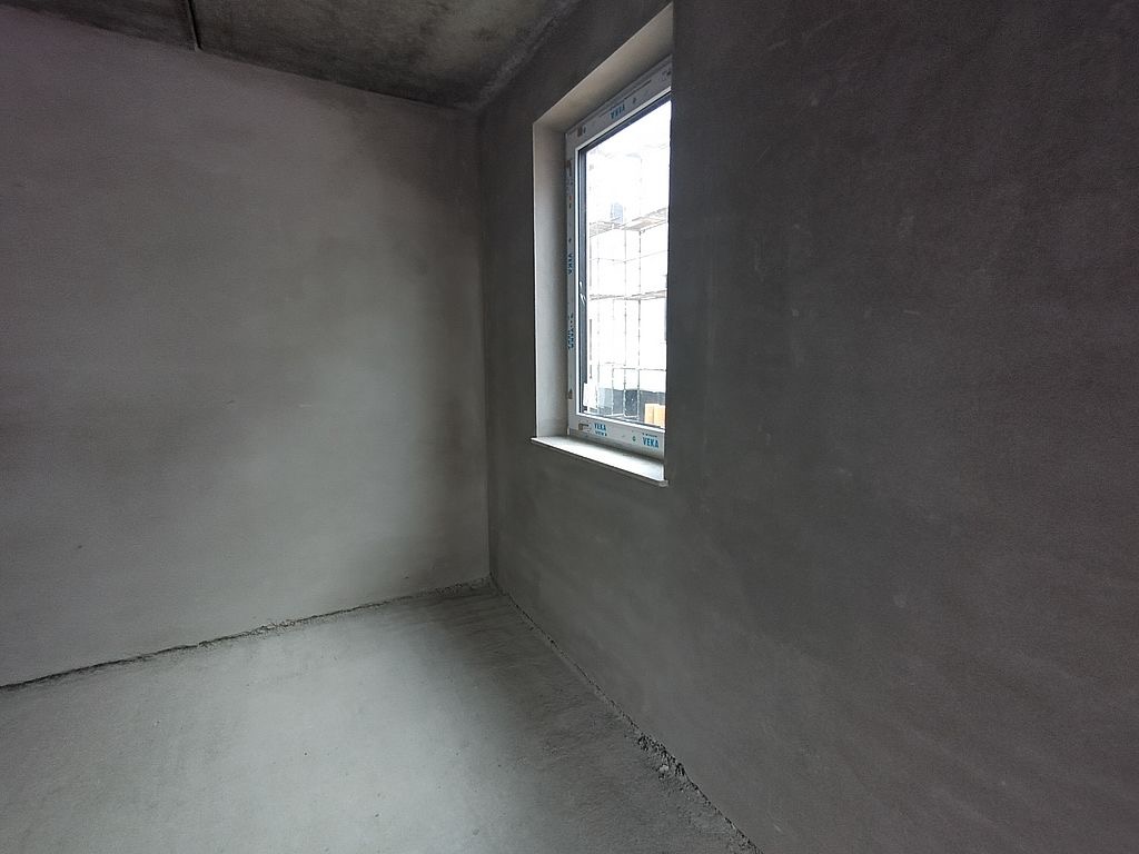Surowe wnętrze po tynkowaniu maszynowym, widoczne okno z białymi ramami. Szare ściany i podłoga, przygotowane do dalszych prac wykończeniowych.