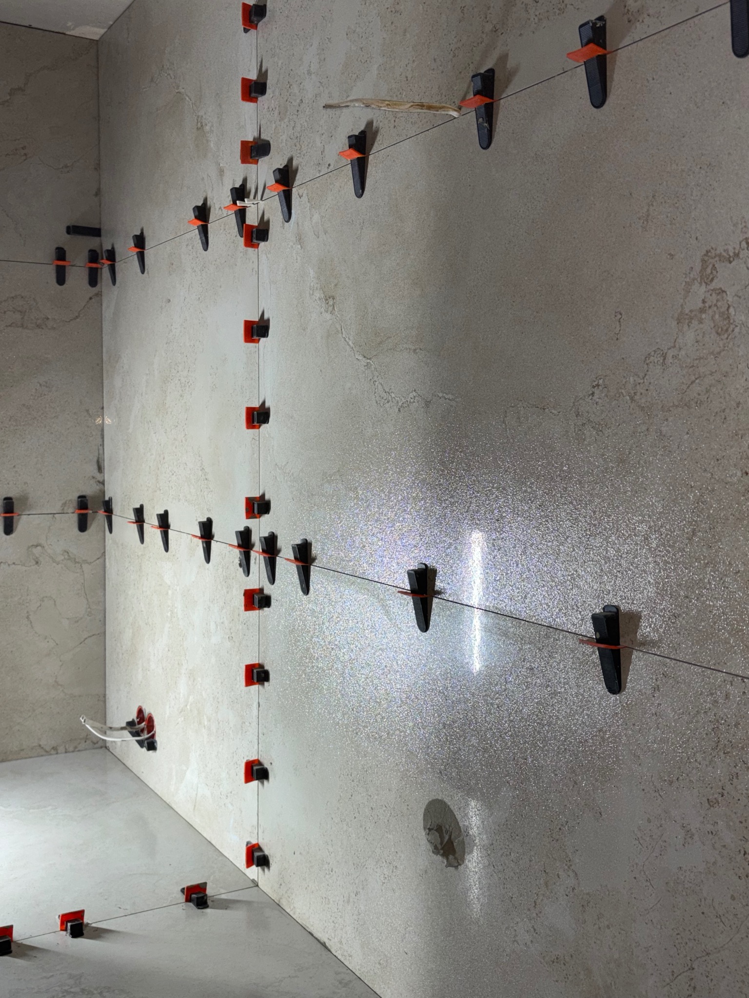 Ściana i podłoga w trakcie układania płytek, z użyciem systemu poziomowania płytek (klipsy i kliny) oraz sznurka wyznaczającego linię. Widoczne detale faktury płytek.