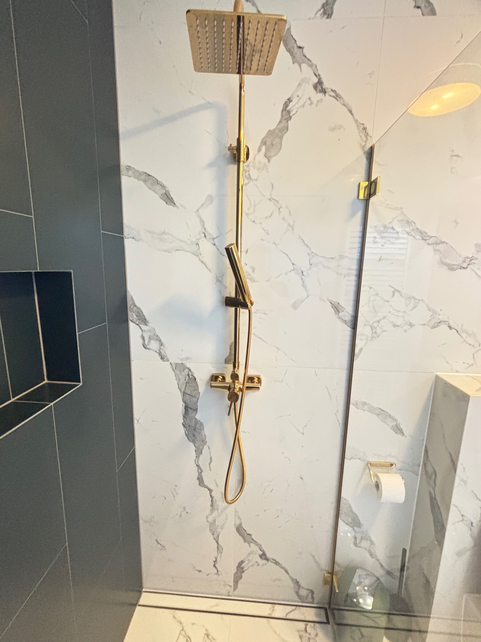 Nowoczesna łazienka z kabiną typu walk-in, wykończona płytkami imitującymi marmur i złotą armaturą, w tym deszczownicą i słuchawką prysznicową.