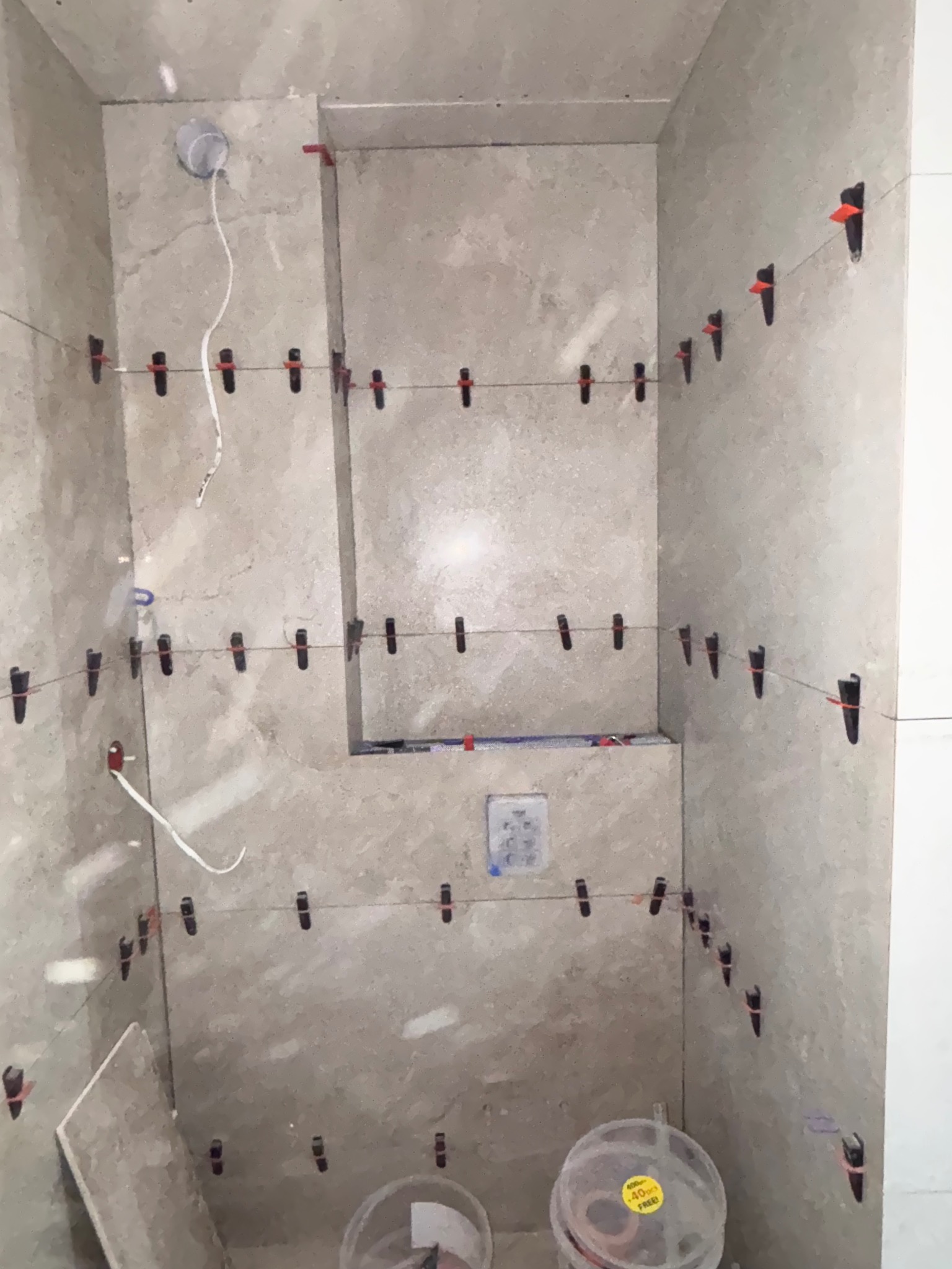 Ściany łazienki wyłożone płytkami w trakcie montażu, z użyciem systemu poziomowania płytek. Widoczne wiaderka i fragment płytki na podłodze.