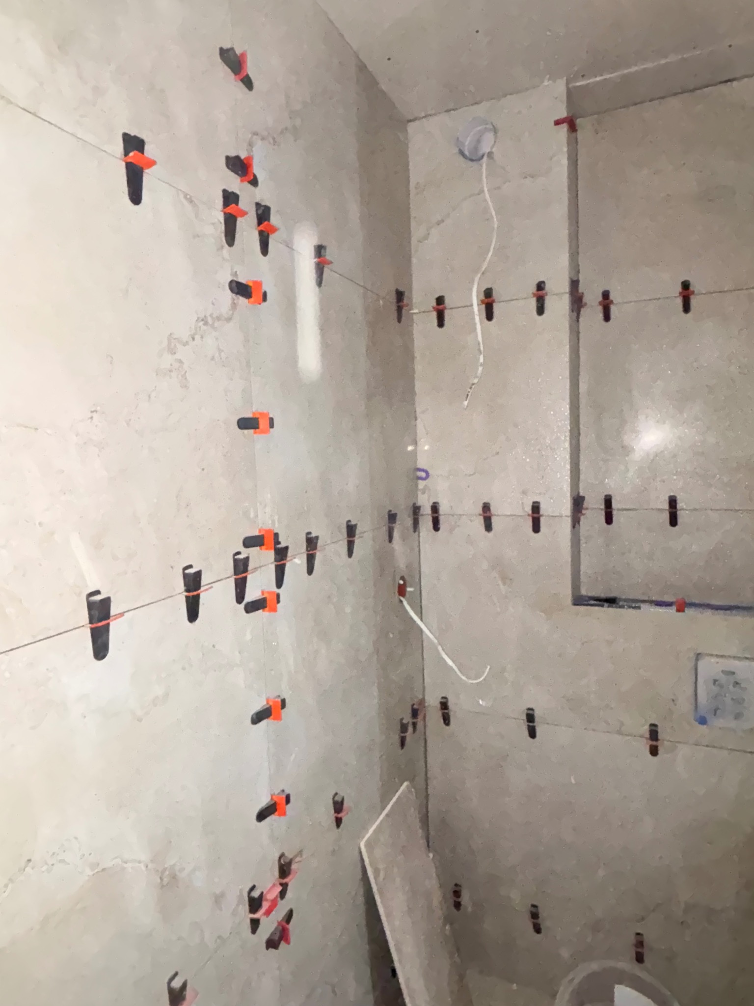 Ściana łazienki w trakcie układania płytek, z systemem poziomowania (klipsy i kliny) oraz widocznymi przewodami elektrycznymi. Jasne płytki imitujące kamień.