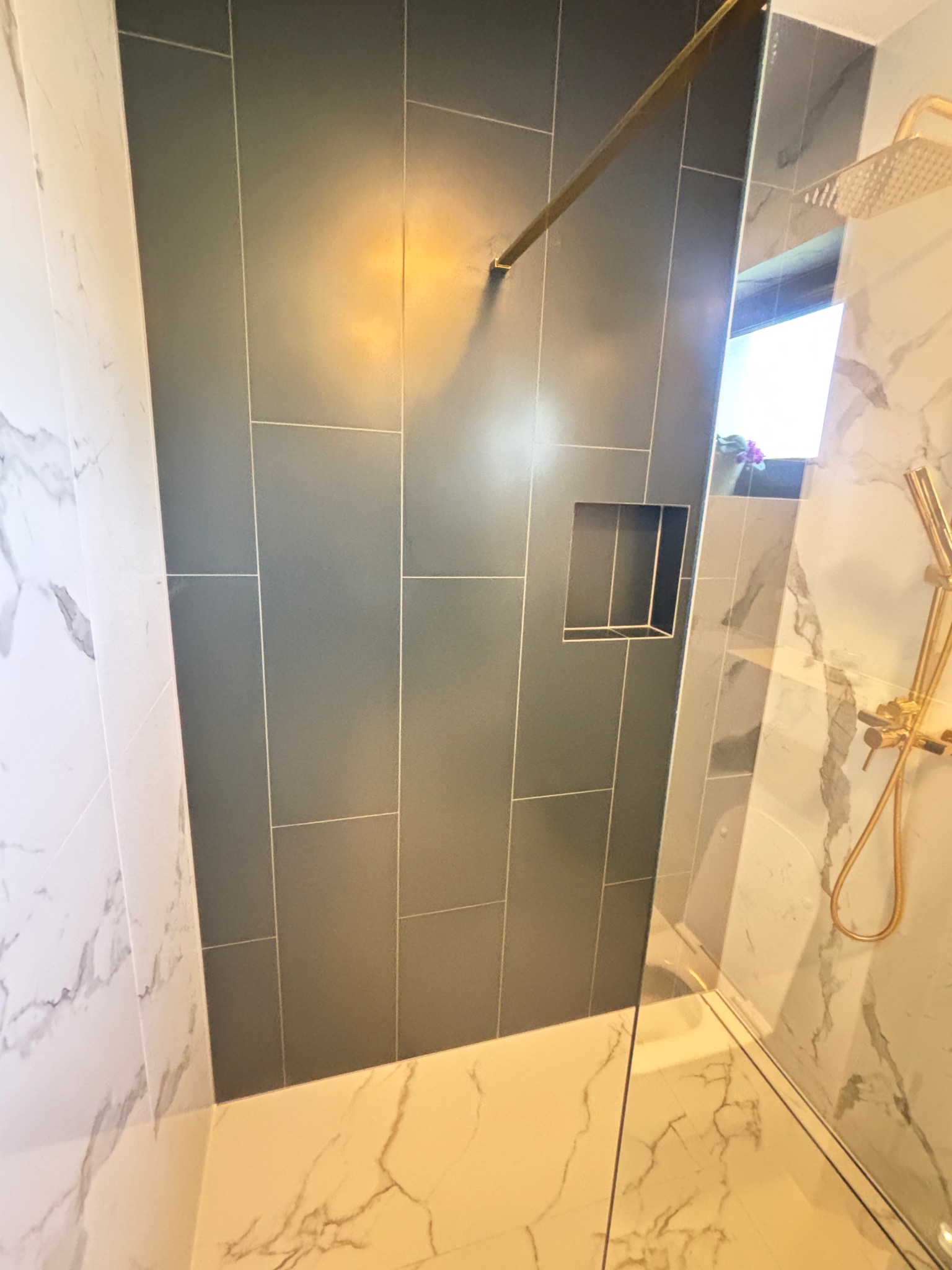 Nowoczesna kabina prysznicowa z szarymi płytkami i wnęką, wykończona marmurem i złotą armaturą. Minimalistyczny design z dbałością o detale.