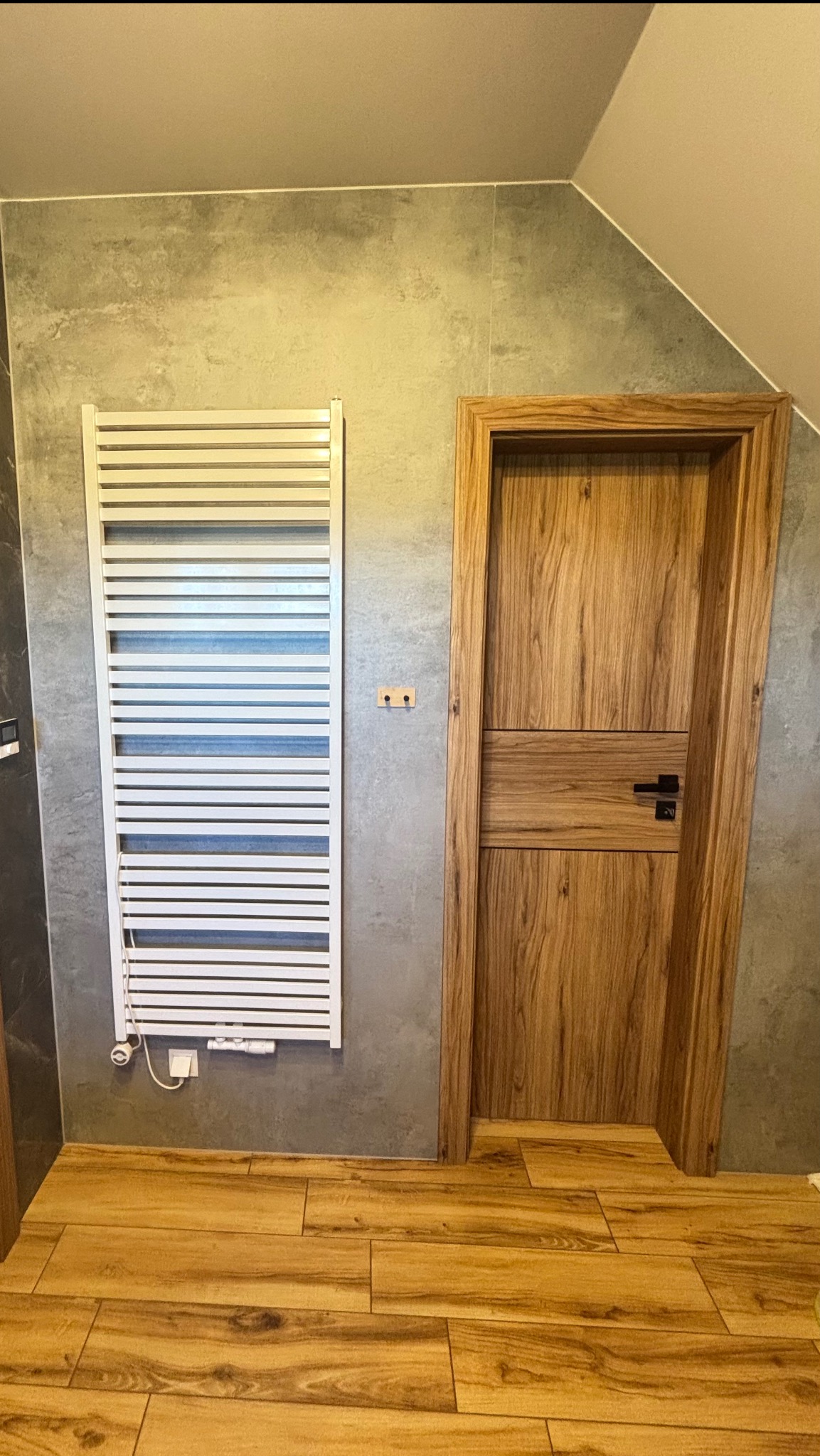 Wykończona łazienka z grzejnikiem drabinkowym, drewnianą podłogą i drzwiami z drewnianą ościeżnicą. Szare płytki na ścianach. Ukośny sufit.