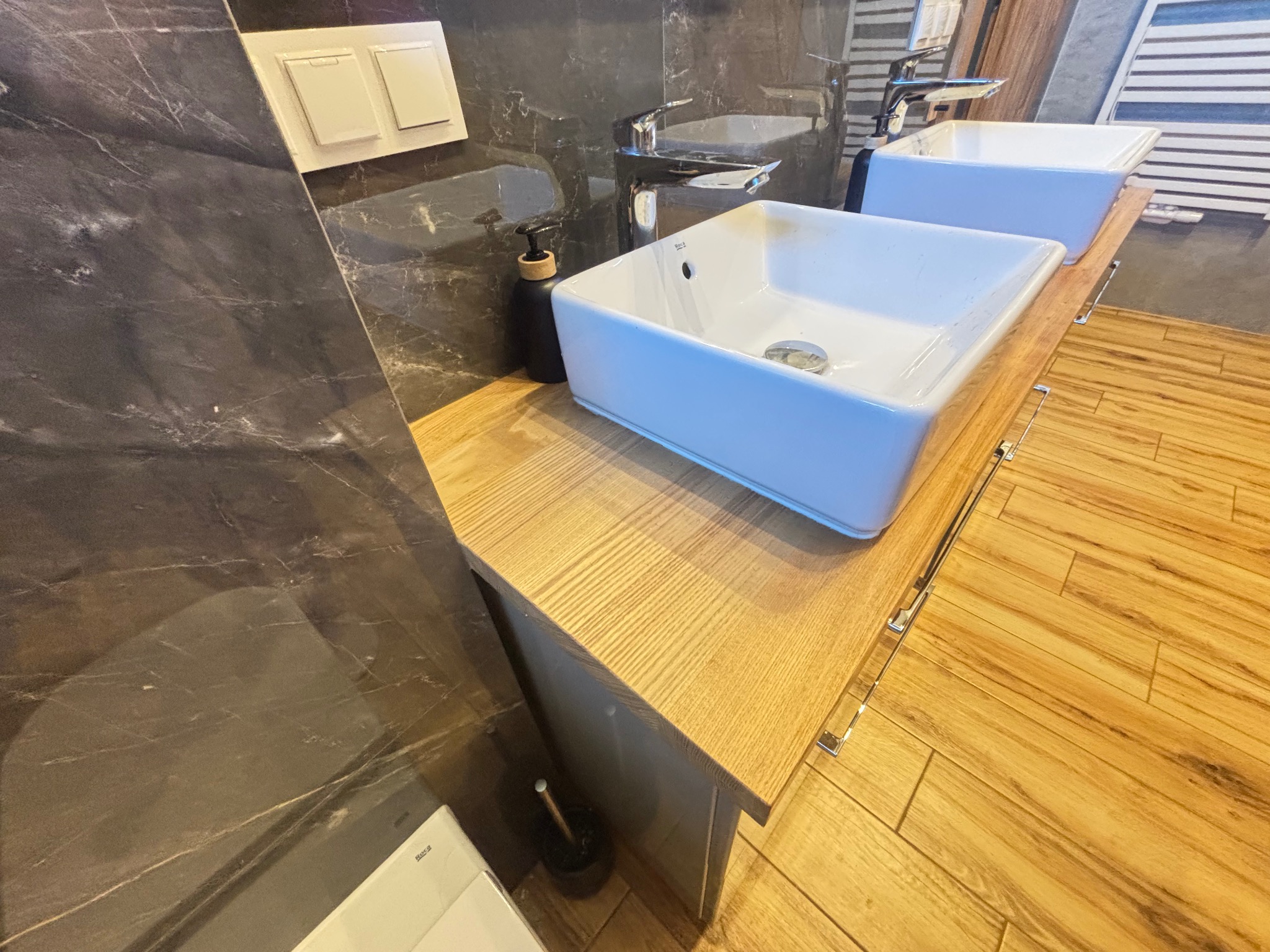 Nowoczesna łazienka z dwiema białymi umywalkami nablatowymi na drewnianym blacie. Ciemne, marmurowe płytki na ścianie i drewniana podłoga. Minimalistyczny design.