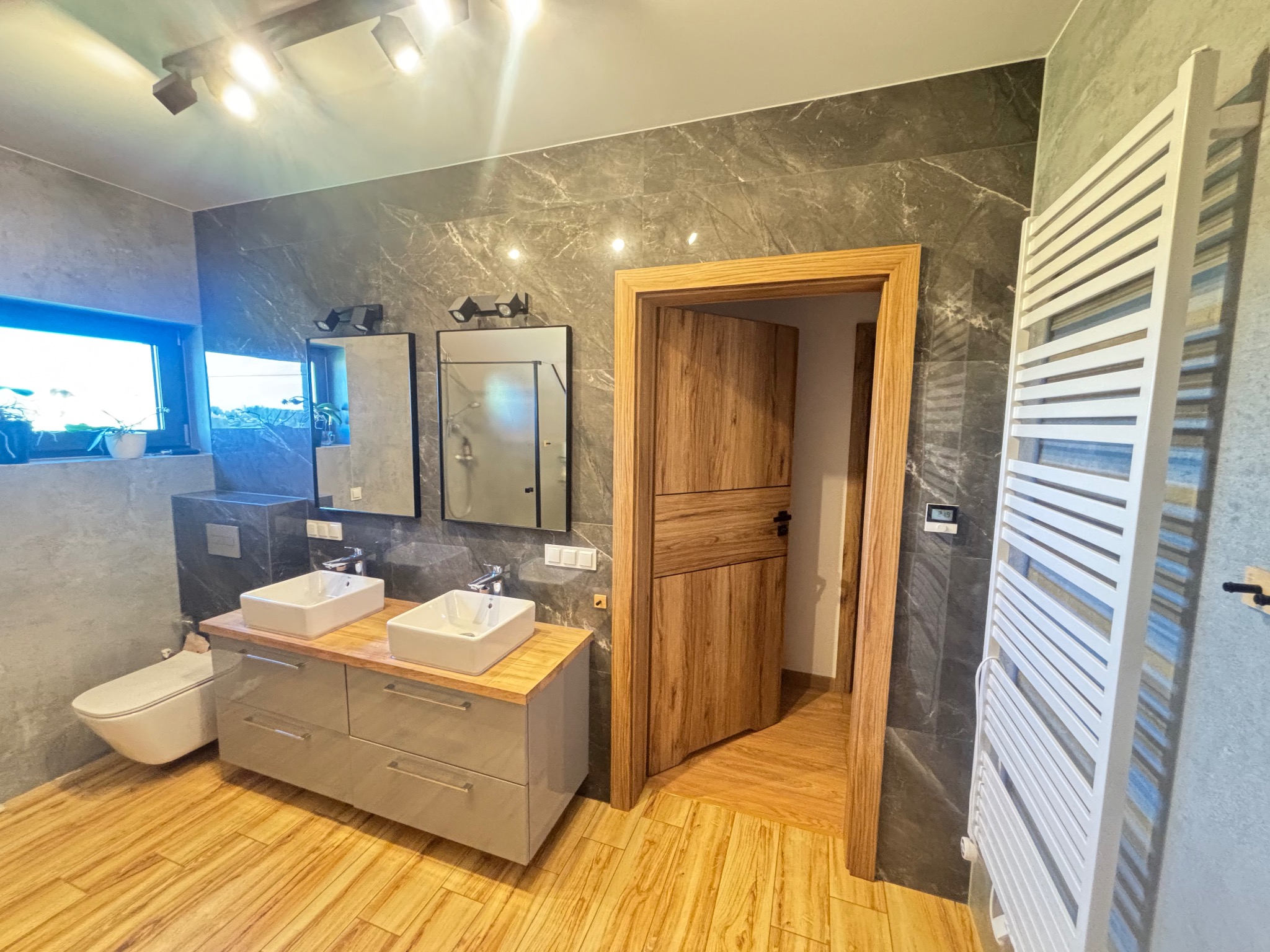 Nowoczesna łazienka z drewnianą podłogą i drzwiami, szare płytki imitujące marmur na ścianach, biała drabinka grzejnikowa i podwójna umywalka.