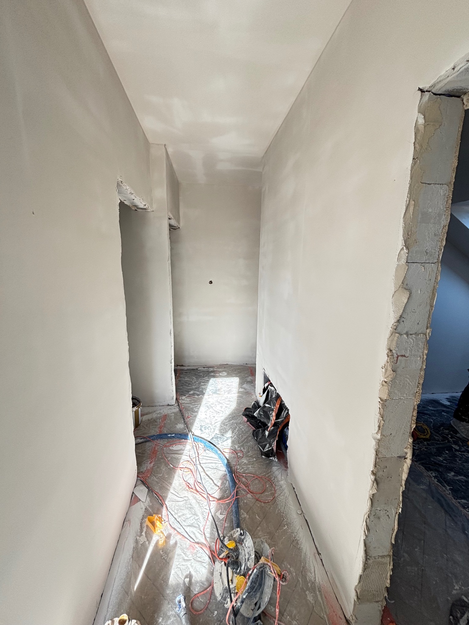 Korytarz w trakcie prac wykończeniowych: świeżo położone gładzie na ścianach, widoczne narzędzia i kable na podłodze, surowy fragment ściany przy wejściu.