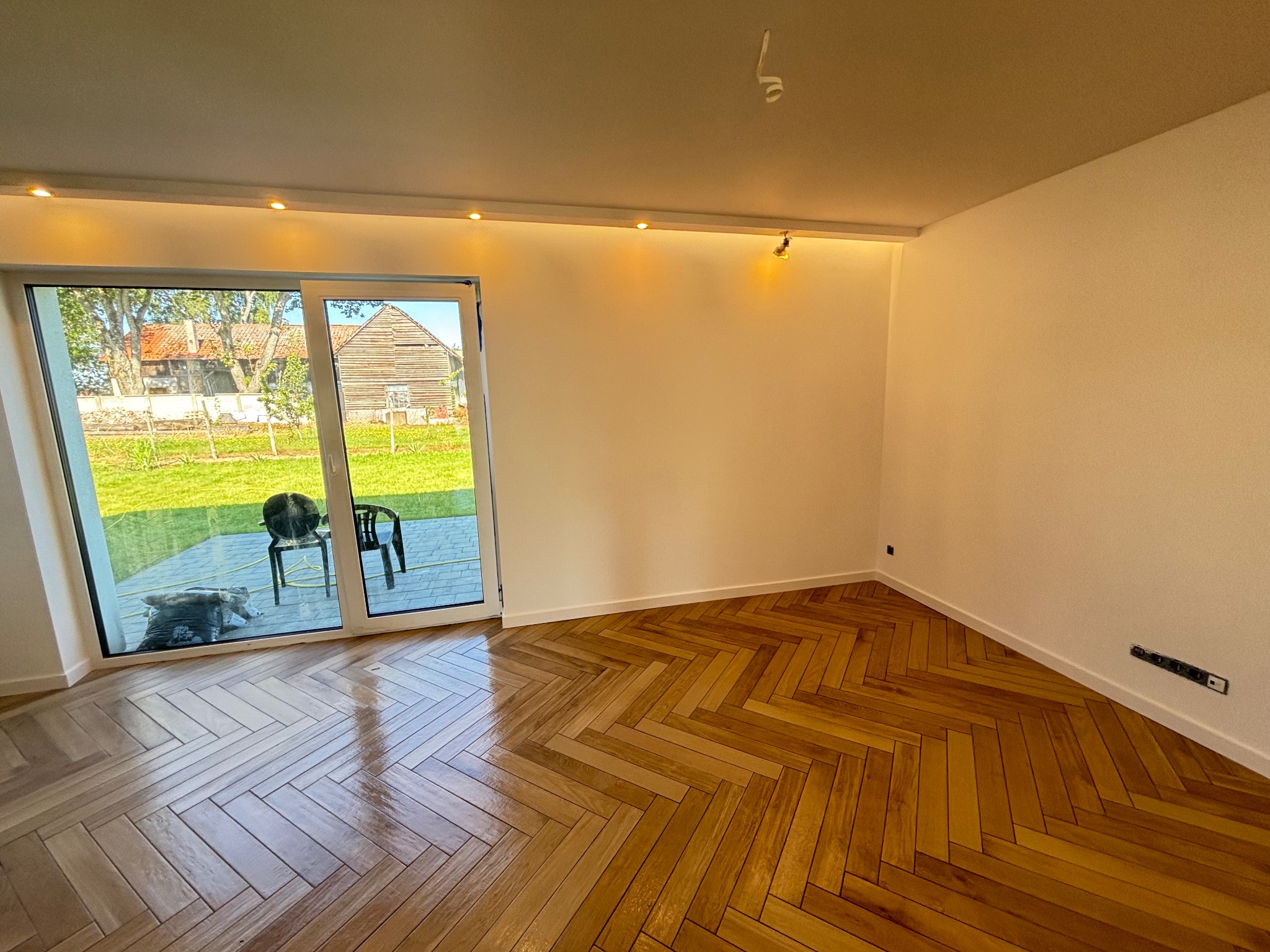 Jasne, odnowione wnętrze z parkietem w jodełkę, białymi ścianami i dużym oknem tarasowym z widokiem na zieleń. Nowoczesne oświetlenie sufitowe.