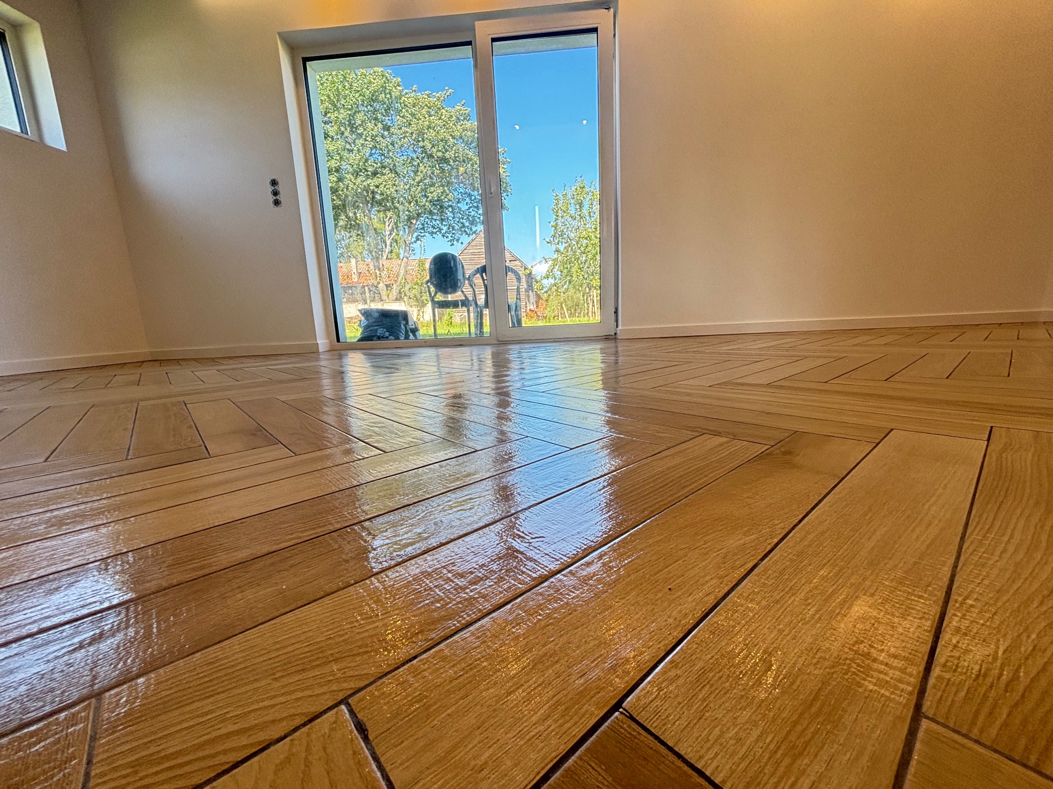 Połyskująca, drewnopodobna podłoga ułożona w jodełkę, z widokiem na zieleń przez przesuwne drzwi balkonowe. Minimalistyczne wnętrze z jasnymi ścianami.