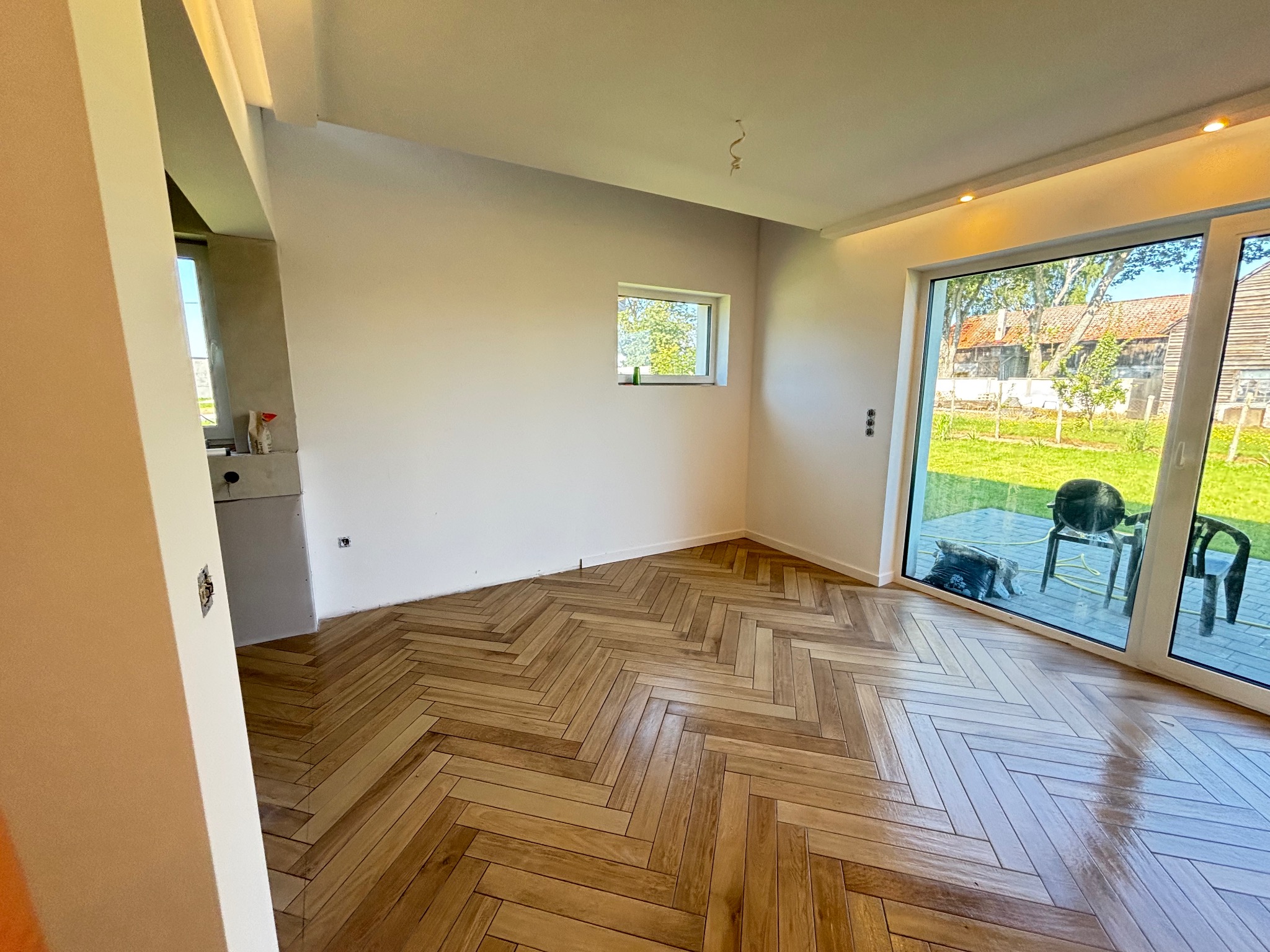 Jasne, parkietowe panele ułożone w jodełkę w przestronnym, odnowionym pokoju z dużym oknem tarasowym i świeżo pomalowanymi ścianami. Widok na zieleń za oknem.