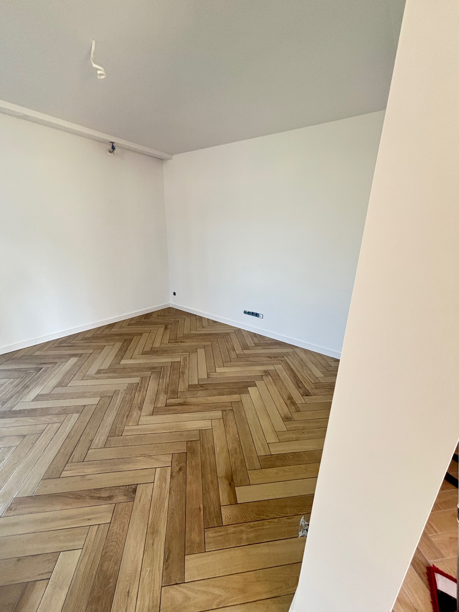 Jasne, puste wnętrze z parkietem ułożonym w jodełkę. Świeżo pomalowane ściany i listwy przypodłogowe. Widoczny fragment kolumny po prawej stronie kadru.