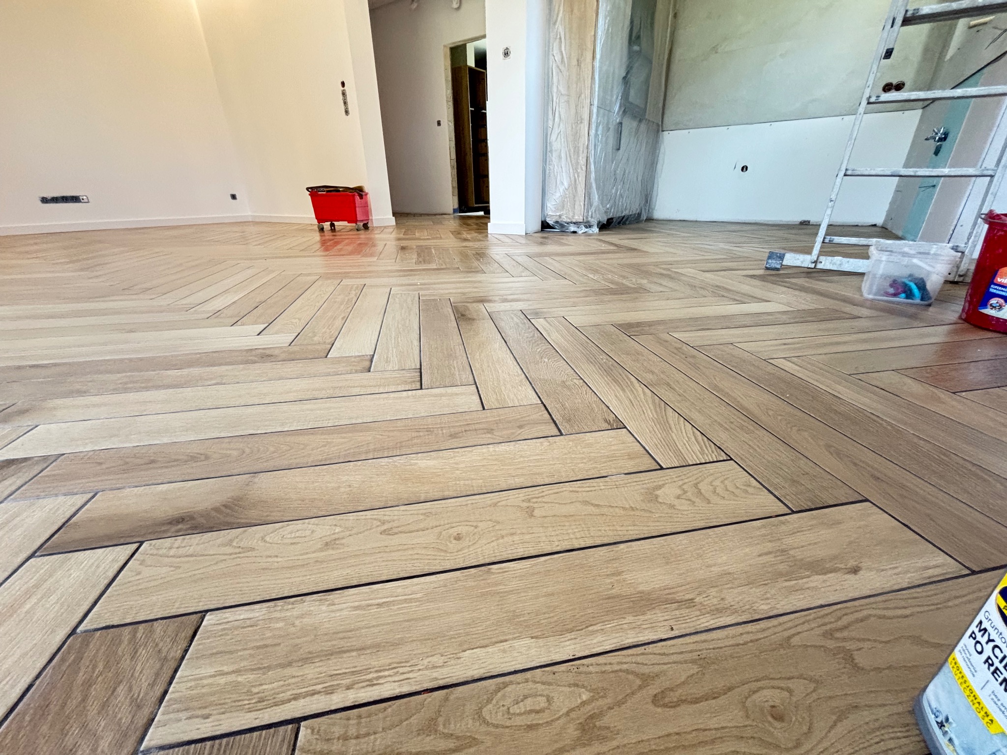 Ukończona podłoga z płytek imitujących drewno, ułożonych w jodełkę. Widoczne narzędzia i akcesoria malarskie, sugerujące świeżo zakończony remont. Jasne wnętrze.