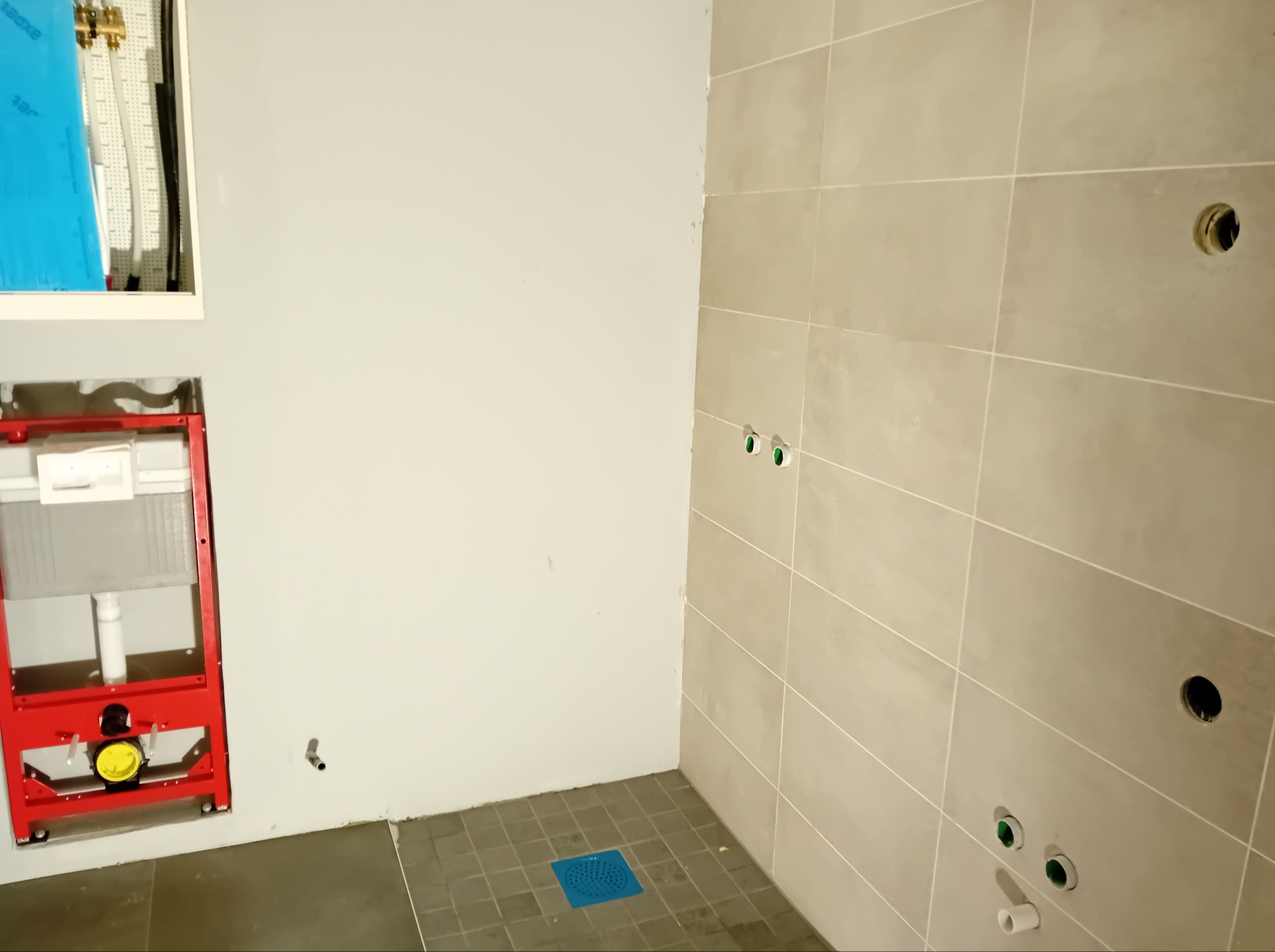 Łazienka w trakcie prac instalacyjnych i okładzinowych: zamontowany czerwony stelaż podtynkowy WC, beżowe płytki ścienne z otworami na armaturę oraz ciemne płytki podłogowe z niebieskim odpływem.