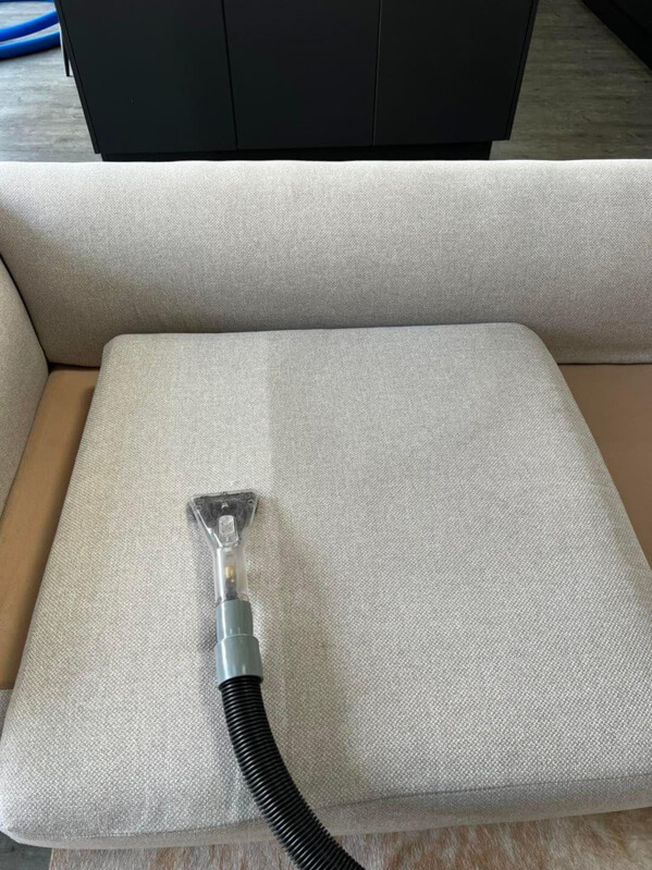 Pranie jasnej tapicerki sofy specjalistycznym odkurzaczem. Widoczna różnica w czystości po lewej stronie, ciemne meble w tle. Efekt czyszczenia na siedzisku.