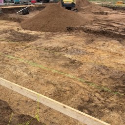 PT-BUD Piotr Tęcza - Przygotowanie terenu pod budowę: wykopane fundamenty, drewniane szalunki, mini koparka i zaparkowany bus na działce. Widok na domy w tle.