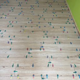 PT-BUD Piotr Tęcza - Układanie podłogi z płytek imitujących drewno, z użyciem kolorowych klipsów poziomujących. Widoczne fragmenty ścian w odcieniach beżu i zieleni.