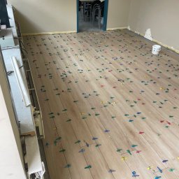 PT-BUD Piotr Tęcza - Układanie paneli podłogowych z użyciem kolorowych klipsów poziomujących w jasnym pomieszczeniu biurowym. Widoczne listwy przypodłogowe i grzejnik.