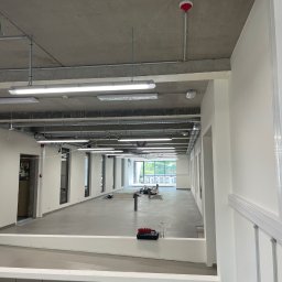 PT-BUD Piotr Tęcza - Wnętrze biura w trakcie prac wykończeniowych: szara posadzka, białe ściany, surowy betonowy sufit z instalacjami. Widoczne narzędzia i materiały budowlane w oddali.