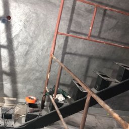 PT-BUD Piotr Tęcza - Ściana wykończona w szarym odcieniu, z widocznym rusztowaniem malarskim i narzędziami. Widać przygotowania do dalszych prac wykończeniowych.