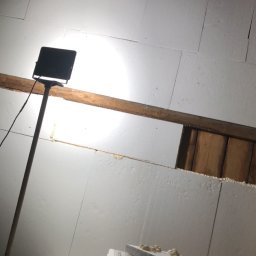 PT-BUD Piotr Tęcza - Montaż płyt styropianowych na ścianie z drewnianą belką. Widoczna lampa oświetlająca miejsce pracy i narzędzia na styropianie. Etap ocieplania pomieszczenia.