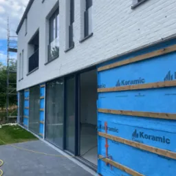 Montaż elewacji budynku z widoczną warstwą izolacyjną (niebieska membrana) i drewnianymi łatami. Nowoczesna architektura, biała cegła, duże okna przesuwne.