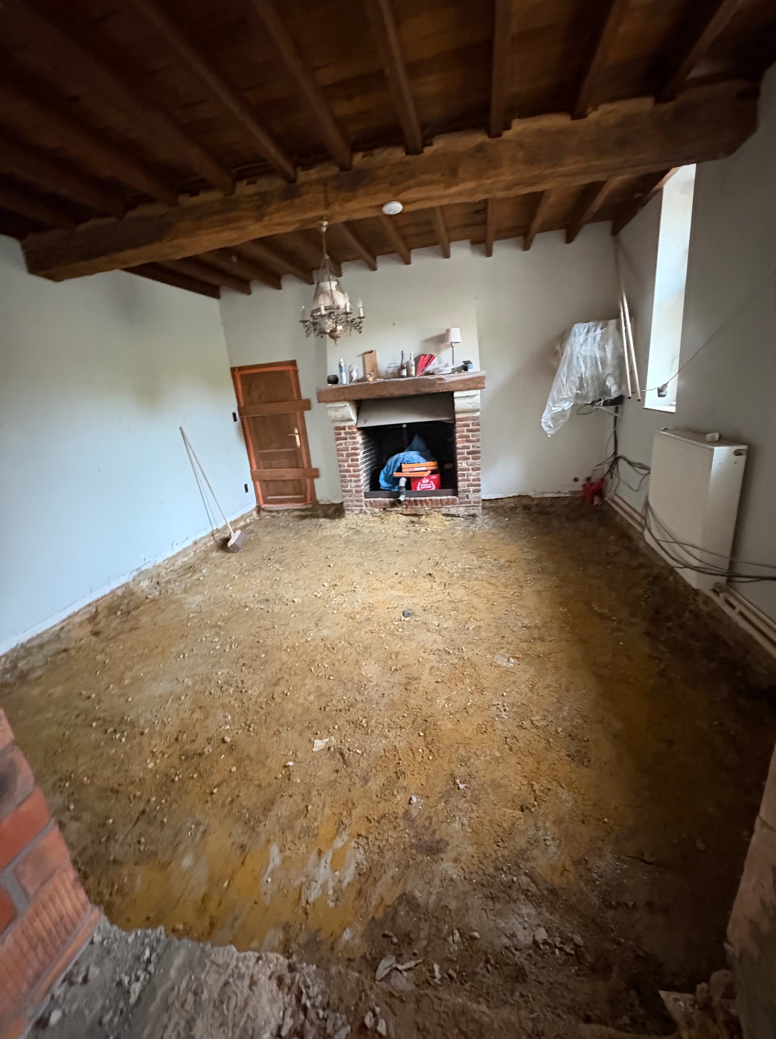 Pokój w trakcie remontu: odsłonięta podłoga, ceglany kominek, drewniany sufit z belkami i żyrandolem. Widoczne ślady prac remontowych, sprzęt i narzędzia.