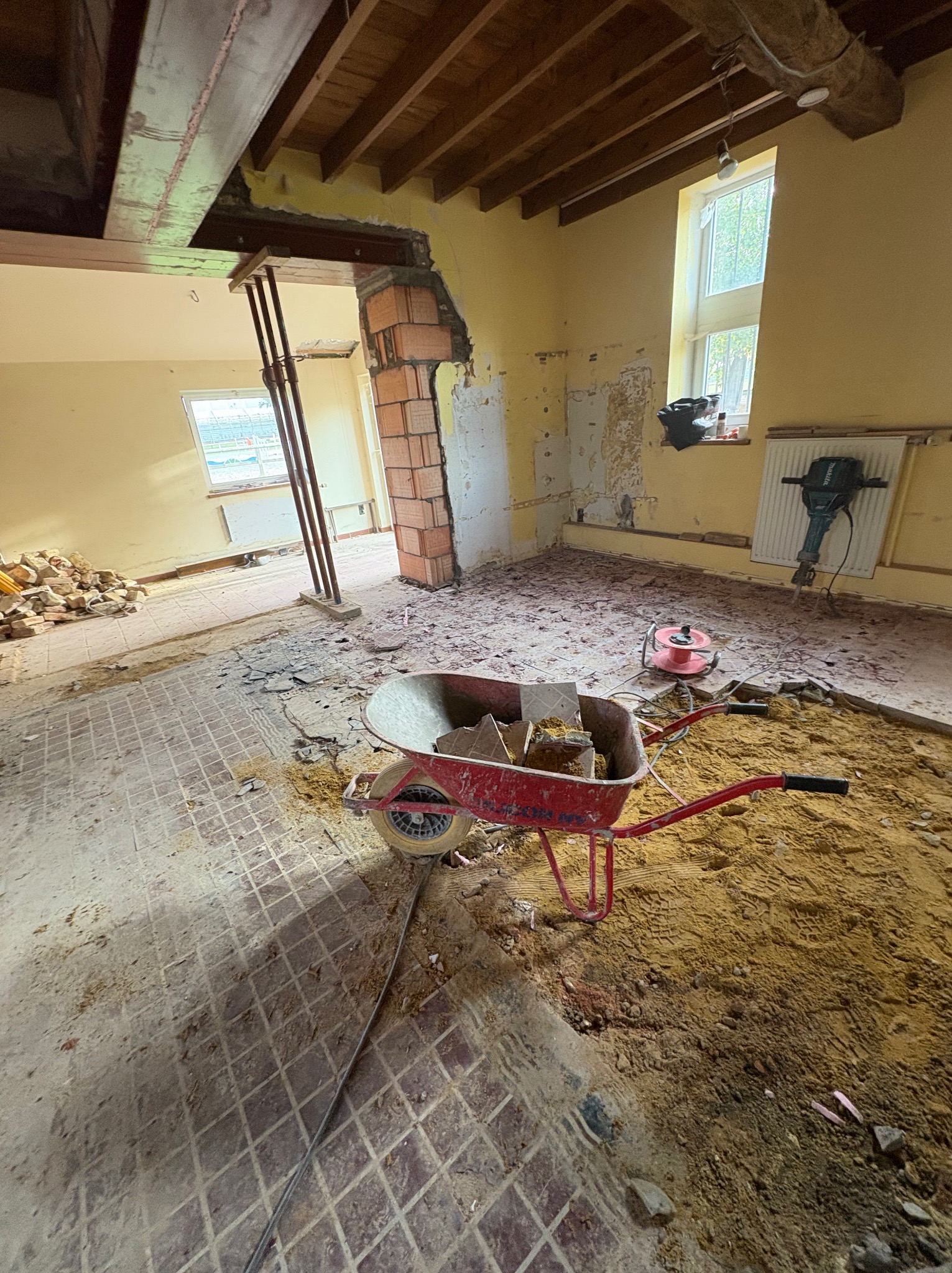 Wnętrze w trakcie remontu: gruz, cegły, taczka z piaskiem, fragment podłogi z wzorem kraty, ściany z resztkami farby i okno. Widoczne elementy konstrukcyjne.